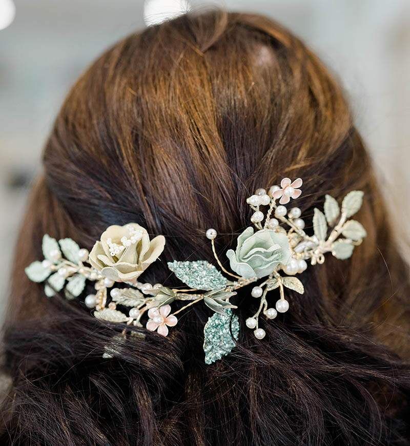 Woman's brown hair styled with floral hair accessory. Hairpiece features cream and green flowers, pearls.