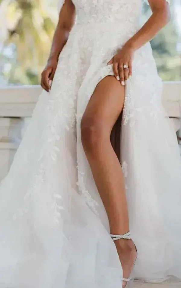 Bride in a white gown with a high slit, showing her leg and ankle strap heels. Outdoors, sunny.