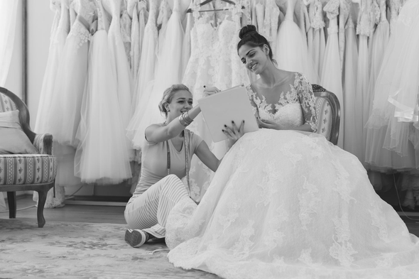 Two women in a bridal shop, one holding a wedding dress, browsing gowns on display.