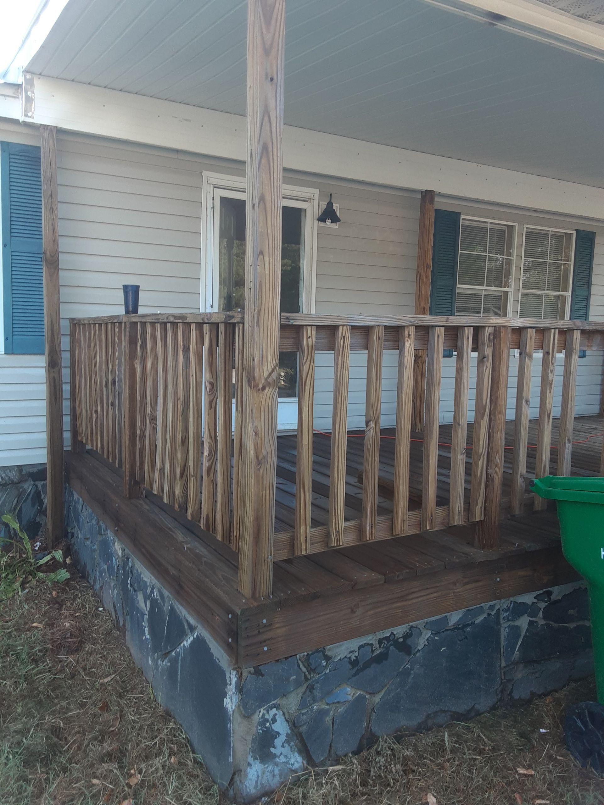 A porch with a wooden railing and a green trash can in front of it.