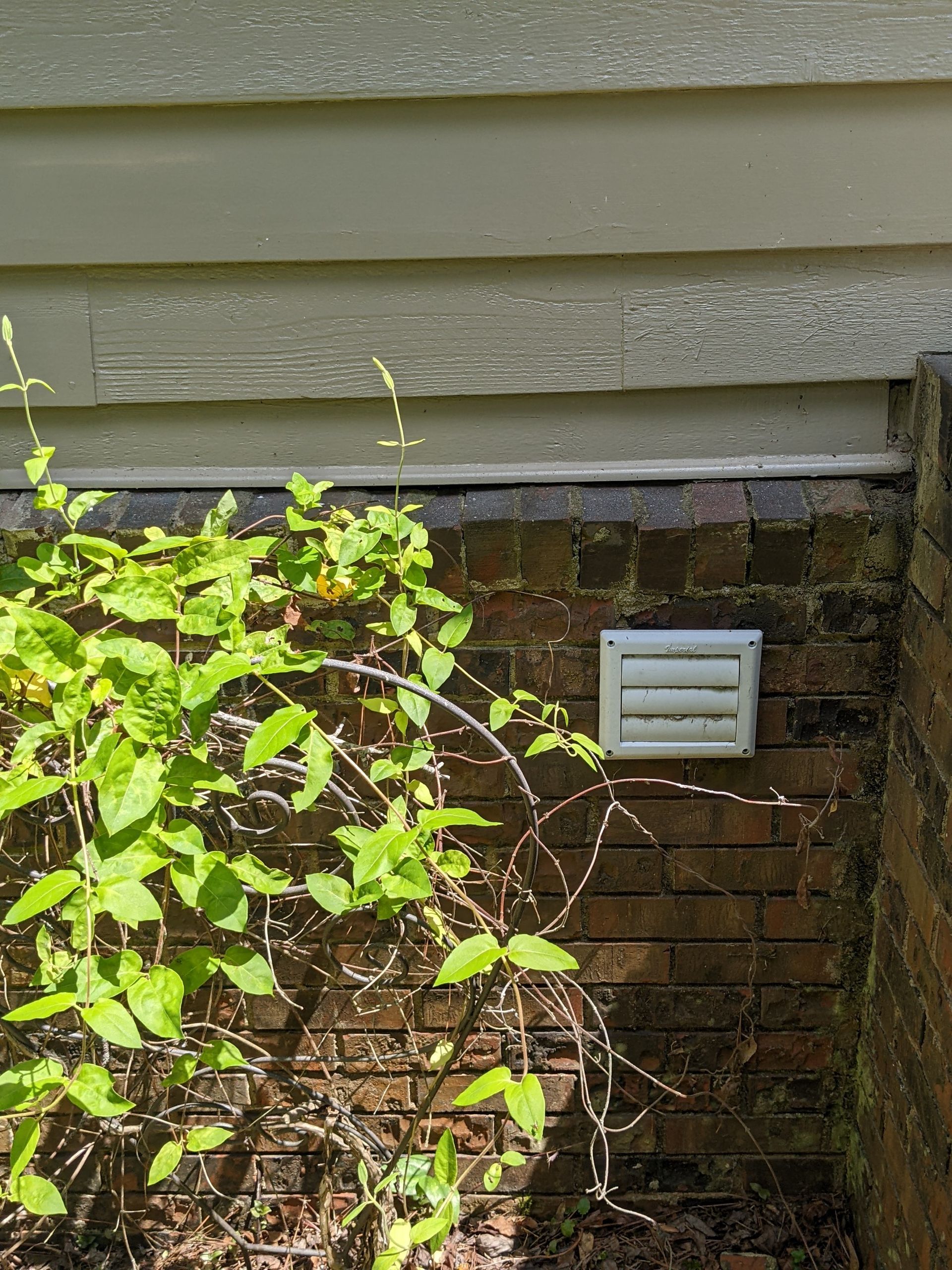 A white vent on a brick wall next to a bush.