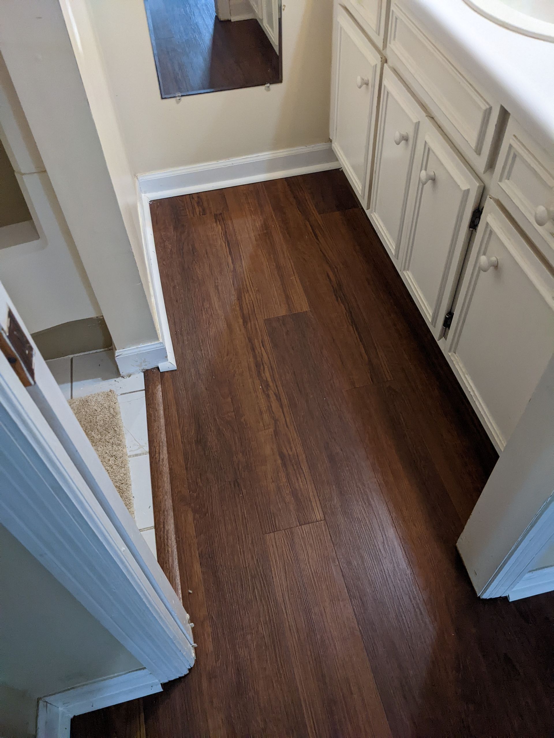 A bathroom with hardwood floors , white cabinets , a sink and a mirror.