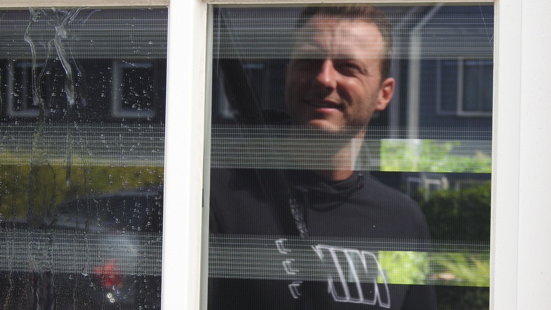 Een man in een zwart shirt kijkt uit een raam.