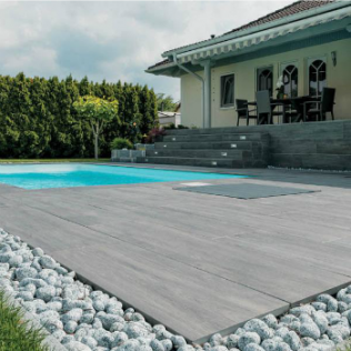 Gray deck surrounds a pool, bordered by white stones and grass, near a beige house with dark furniture.