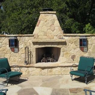 Stone fireplace with two green chairs on a patio. Decorative windows flank the fireplace.
