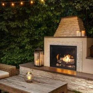 Outdoor fireplace with fire lit, lanterns, and wooden table, surrounded by lush greenery.