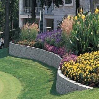 A curved retaining wall with colorful flowers and ornamental grass, beside a green lawn and building.