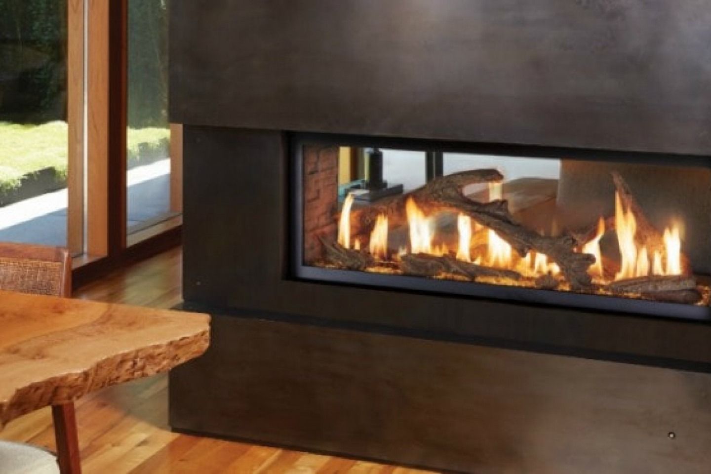 A modern fireplace with glowing flames and logs, framed in dark gray, indoors near a wooden table.
