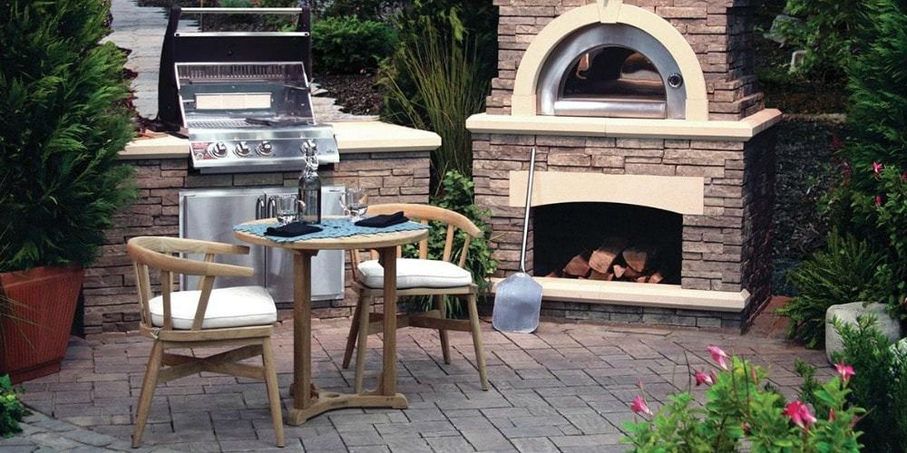 Outdoor patio with grill, pizza oven, table, chairs, and landscaping.