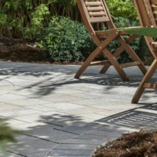 Wooden chairs on a gray stone patio, with shadows and a garden border in background.