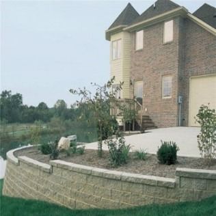 Tan brick house next to a curved retaining wall with landscaping and a small pond in the background.