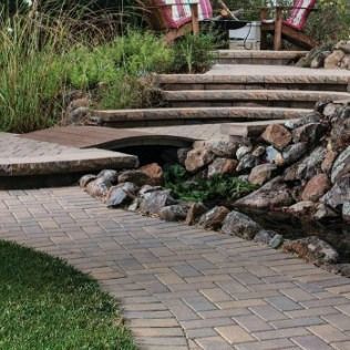 Brick pathway leads to stone steps near a garden with bridge, rocks, and chairs.
