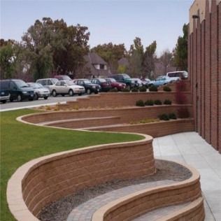 Curving retaining walls with grass and landscaping next to a brick building and a parking lot.