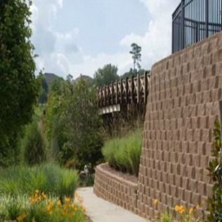 Stone retaining wall and pathway in a garden setting, with green plants and a bridge visible.
