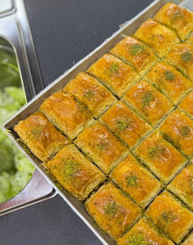 Un vassoio di metallo pieno di pezzi rettangolari di baklava color marrone dorato, ricoperti di pistacchi verdi tritati.