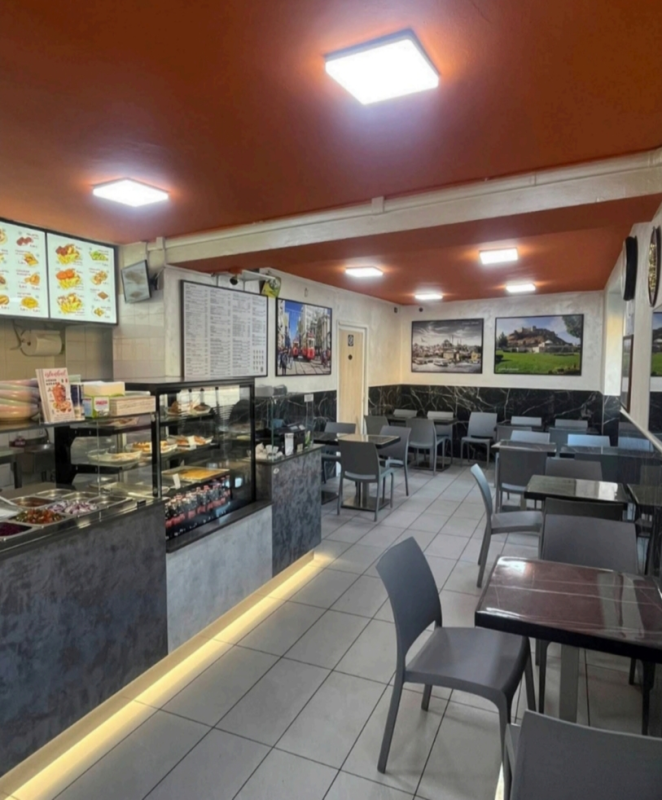 Interno vuoto di una caffetteria con bancone in vetro, lavagne con il menù, tavoli e sedie moderni, illuminati da calde luci a soffitto.