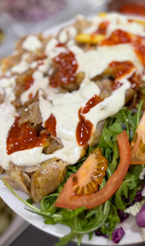 Un piatto di carne di döner kebab ricoperta di salsa bianca all'aglio e salsa rossa al peperoncino, servito su un letto di verdure fresche e pomodoro.