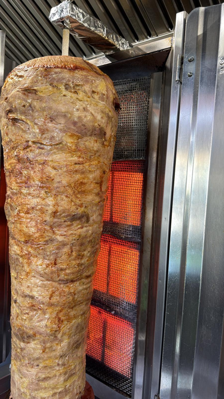 Un alto spiedo verticale, carico di carne arrosto, ruota davanti a elementi riscaldanti arancioni incandescenti.