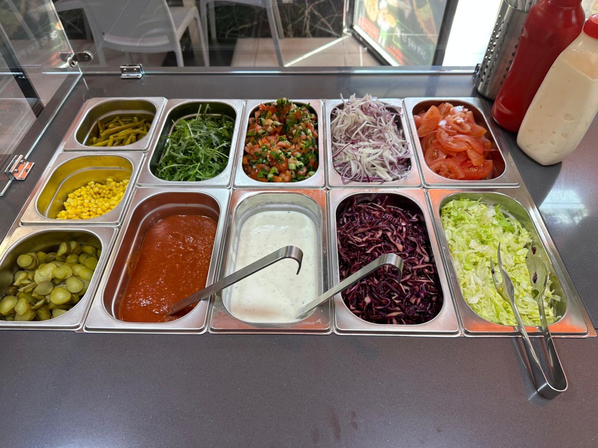 Un'insalateria con vari ingredienti freschi come verdure, salse e condimenti in contenitori metallici rettangolari.