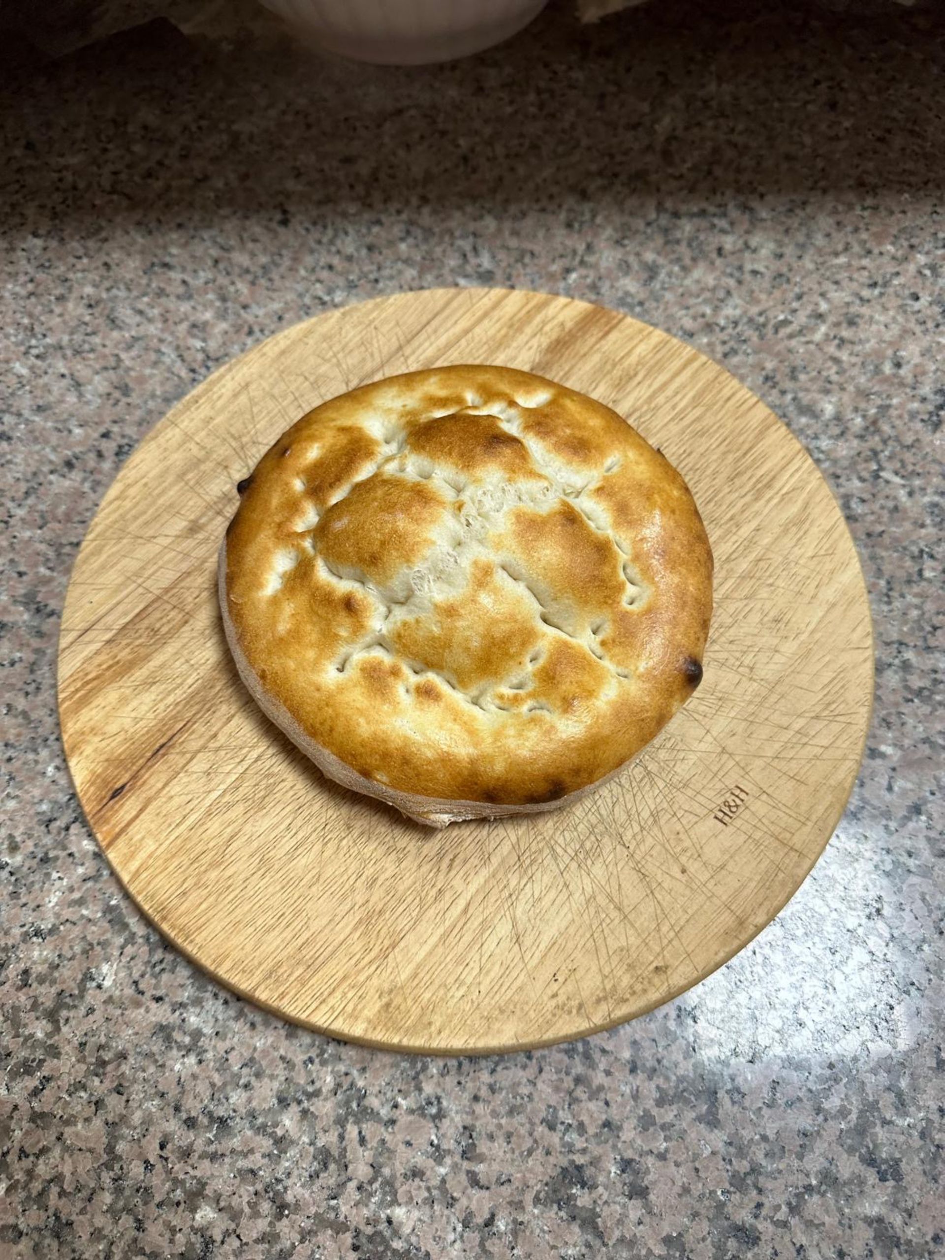 Un panino o biscotto rotondo di colore marrone dorato con la superficie leggermente incavata, posizionato al centro di un piccolo tagliere di legno su un piano di lavoro in granito.
