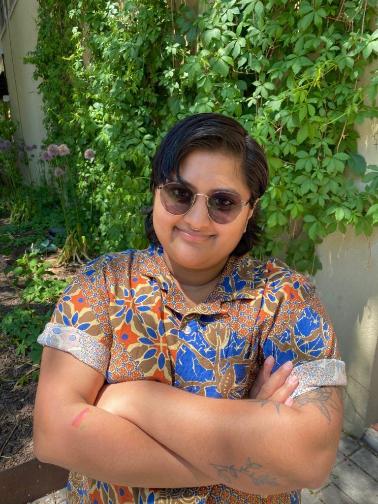 Accessible Futures host Pree Rehal headshot. Pree is a brown person with short dark hair, a nose ring, and tattoos on their arms. They are wearing sun glasses and a brightly printed shirt. They are gently smiling at the camera with their arms crossed and standing infront of a leafy background.