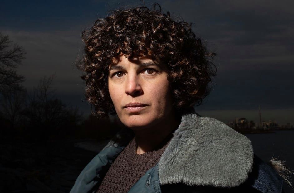 A woman with curly hair is wearing a blue jacket and looking at the camera.