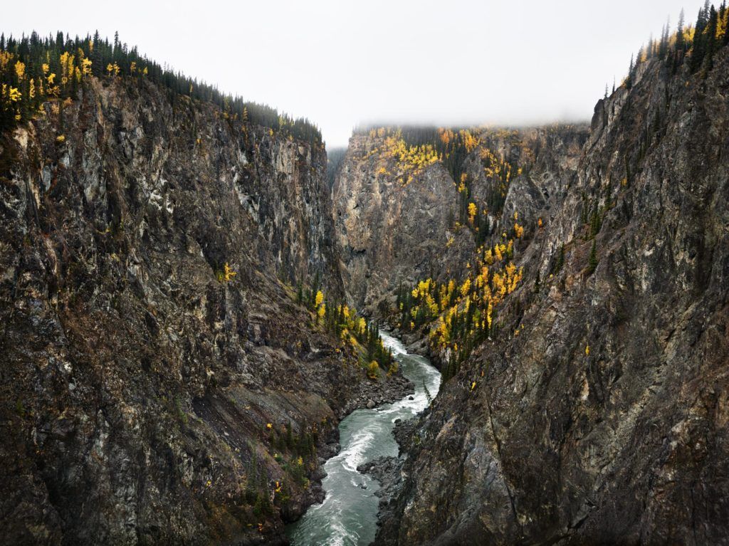 A photo of the Stikine River in Northern British Columbia taken by Edward Burtynsky. A river flows through two rocky mountains. The mountains have slight vegetation on them with yellow coloured trees. Clouds partly cover the top of the mountains. The river appears to be flowing rapidly and strongly in between the mountains.