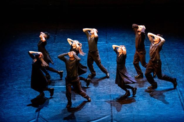 A photo from a stage production of Nuit. Dancers are holding their heads in their hands and looking up towards the ceiling. The dancers are wearing all black and lit up by blue and yellow lights.