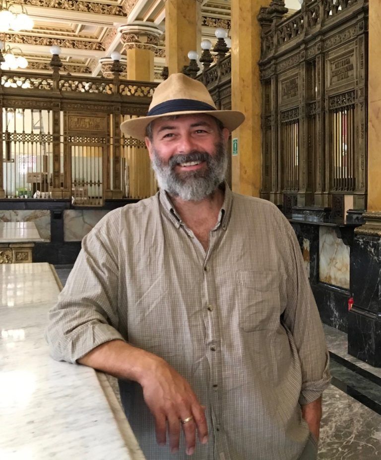 Image Description: Standing in an ornate art nouveau post office, a middle-aged white man with dark brown eyes, a greying beard, and straw fedora faces us and smiles, leaning against a white marble countertop. 