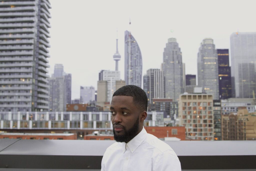 Ian, a Black man with a cropped beard, is wearing a white collared shirt and looking solemnly off to the side. Behind him, we see the city skyline of Toronto on an overcast day.