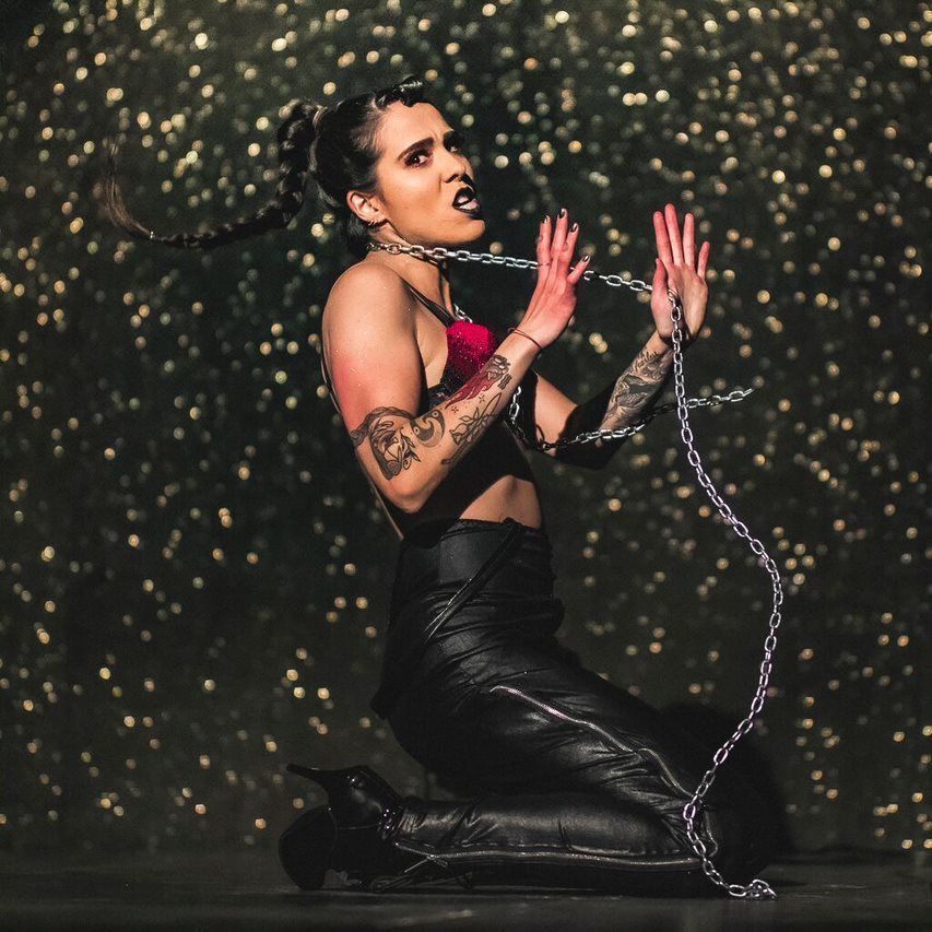 Production photograph of performer Midnight Wolverine, a gender non-conforming Indigenous artist. They are kneeled down on their knees facing the camera in profile with their face towards the camera. A long metal chain is wrapped around their neck and their hands are loosely grasping the chain. The facial expression signifies strength rather than violence. They are wearing black leather pants, black high heel boots, and a red halter top with thin straps. Their hair is long, dark and braided and they are wearing black lipstick and nail polish. The performer is on a stage with a black sparkly backdrop.