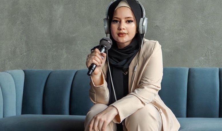 Image Description: This image shows Nisreen sitting on a couch wearing headphones over her head and holding a microphone with her right hand, while she is looking straight at the camera.