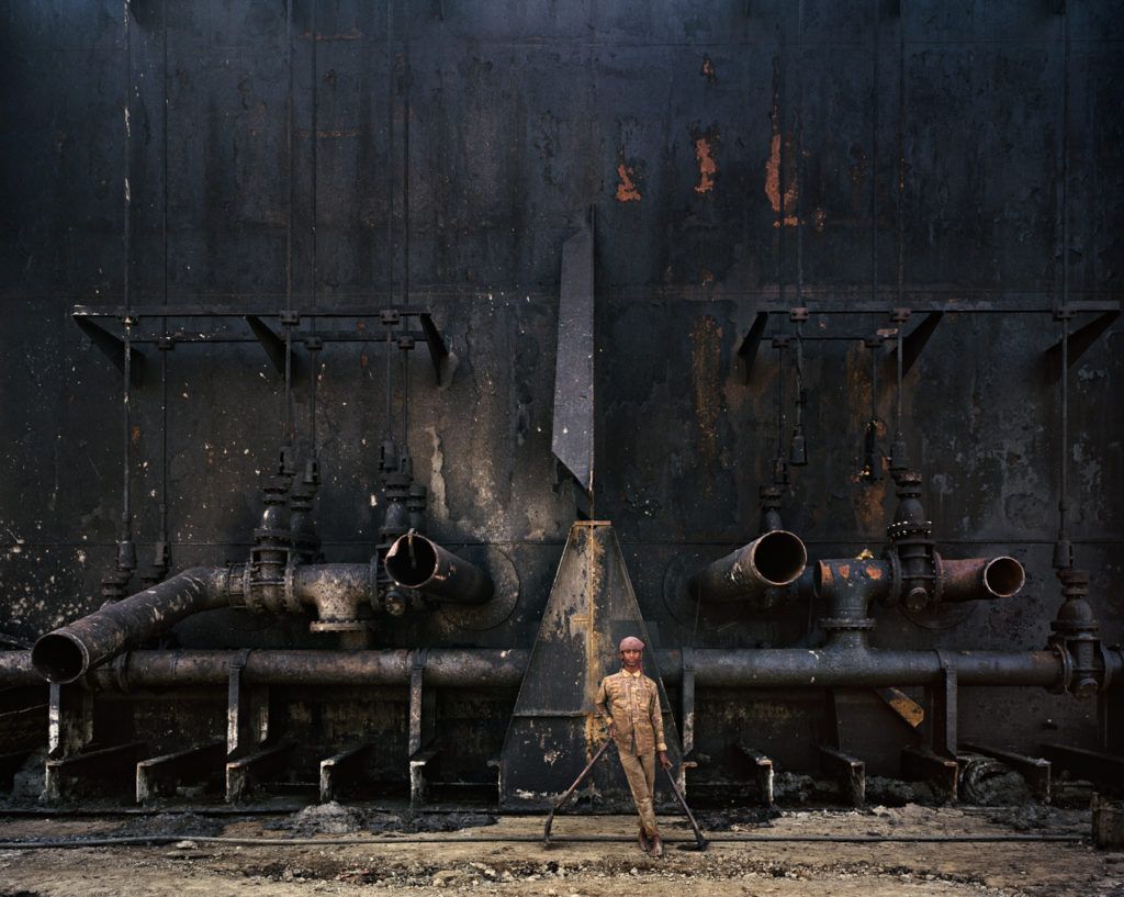 A boy stands in front of a large metal ship. He is working on the process of shipbreaking. He is holding two axes and is wearing a dirty, beige jacket and beige pants. He is barefoot, and his feet are covered with ashes.