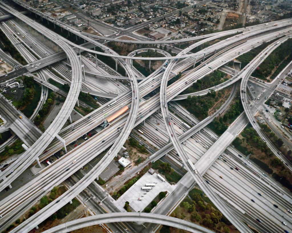 An aerial photo of a highway intersection taken by Edward Burtynsky. This is a bird's eye view of highway lanes winding over each other. Cars and trucks are driving in the many lanes pictured here.