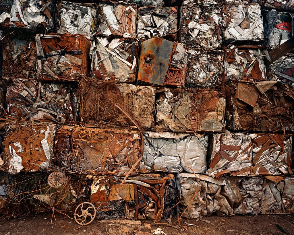 An image of rusty metal molded together, taken by Edward Burtynsky. The scrap metal is tightly packed together in cubes placed on top of each other, forming a rusty wall of metal. 