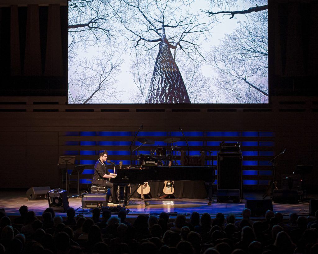 Jeremy Dutcher, a musician, composer, and vocalist is sitting at a grand piano performing at a concert hall. There is a projection behind him of a black and white image of a forest. 