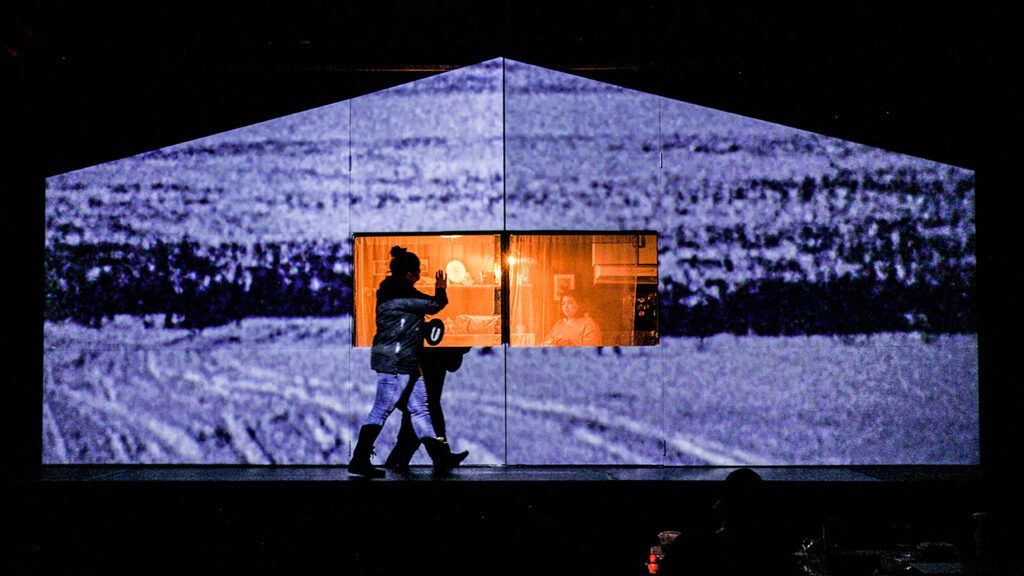 Photo of the stage presentation of Aalaapi. A woman is walking across the stage. On the stage, there is a house with a projection of blue-white snow. There is a window in the house with an orange light glowing out of it.