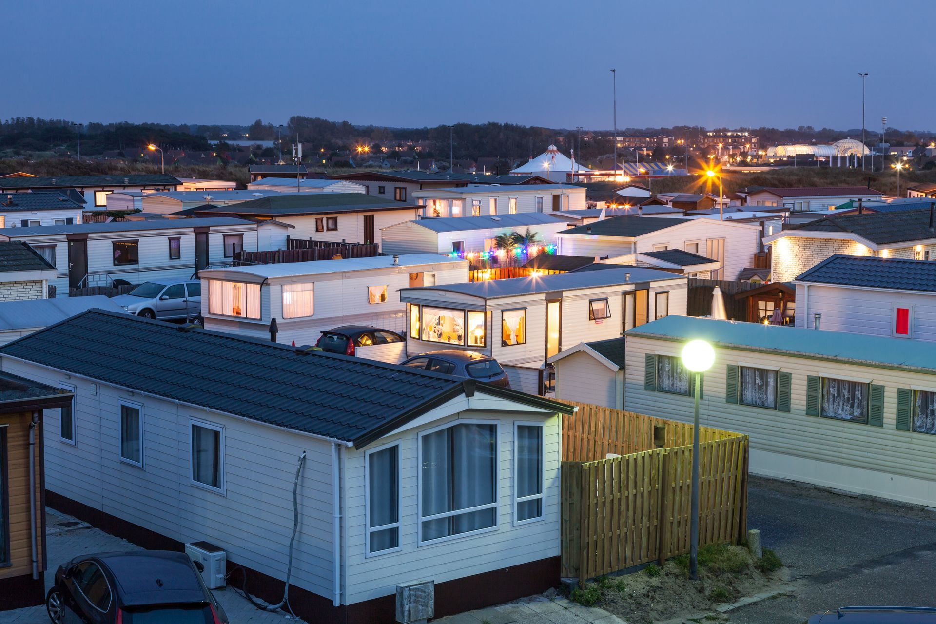 Mobile home community at dusk with lit windows, parked cars, and streetlights across the neighborhood.