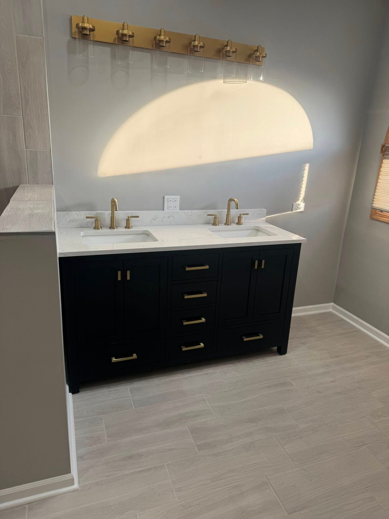 Black double vanity with gold fixtures in a bathroom, under gold vanity lights. Light gray floor.