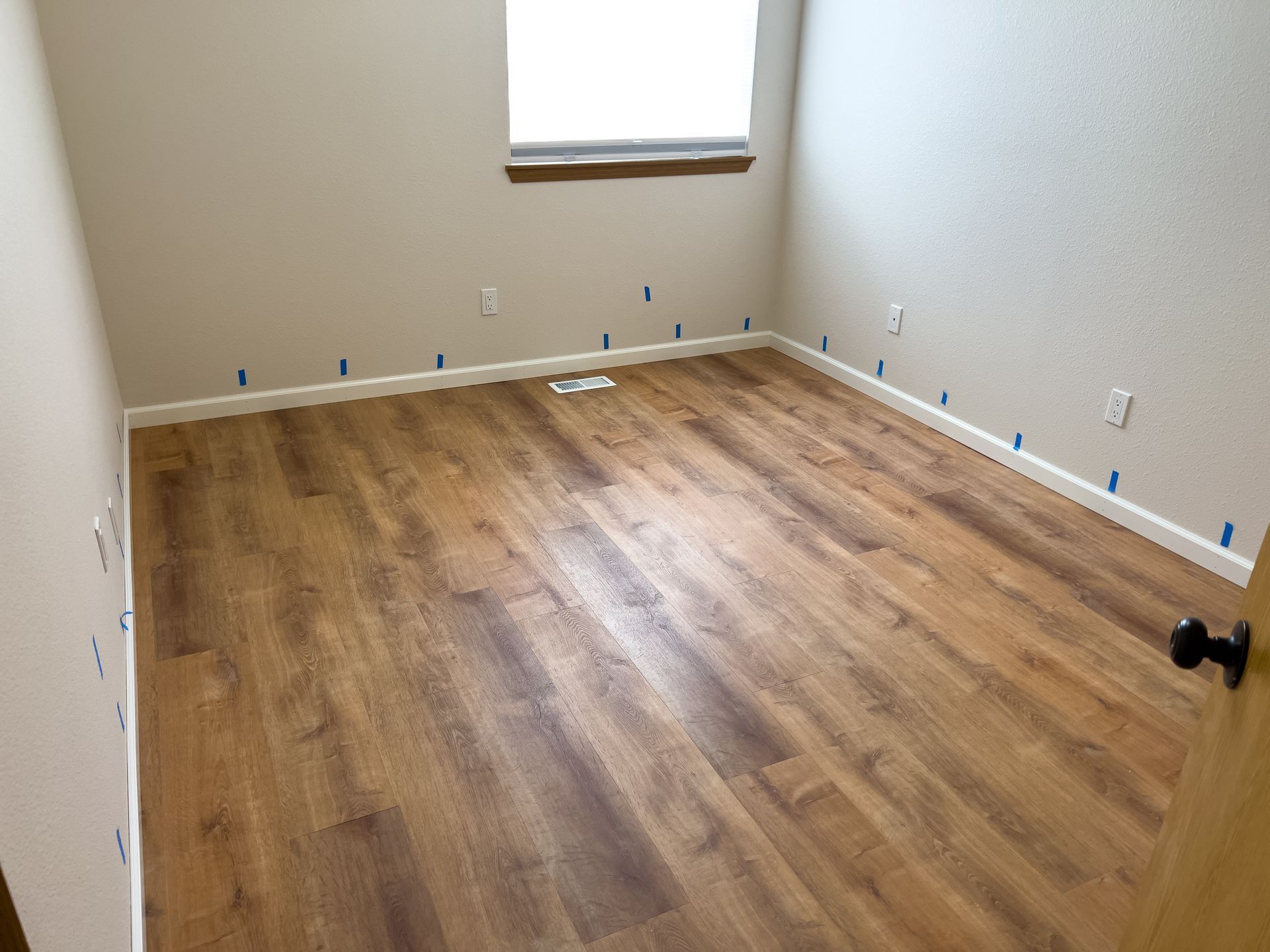 Empty room with new brown wood-look flooring, beige walls, and a window. Blue tape lines the floor edges.