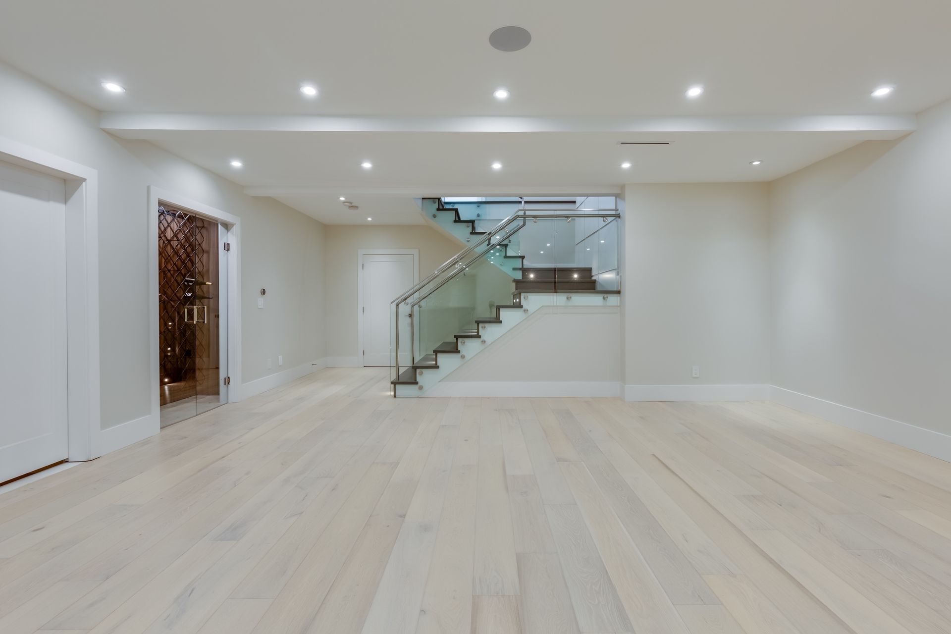 Empty room with white walls and wooden floors, staircase with glass railing.