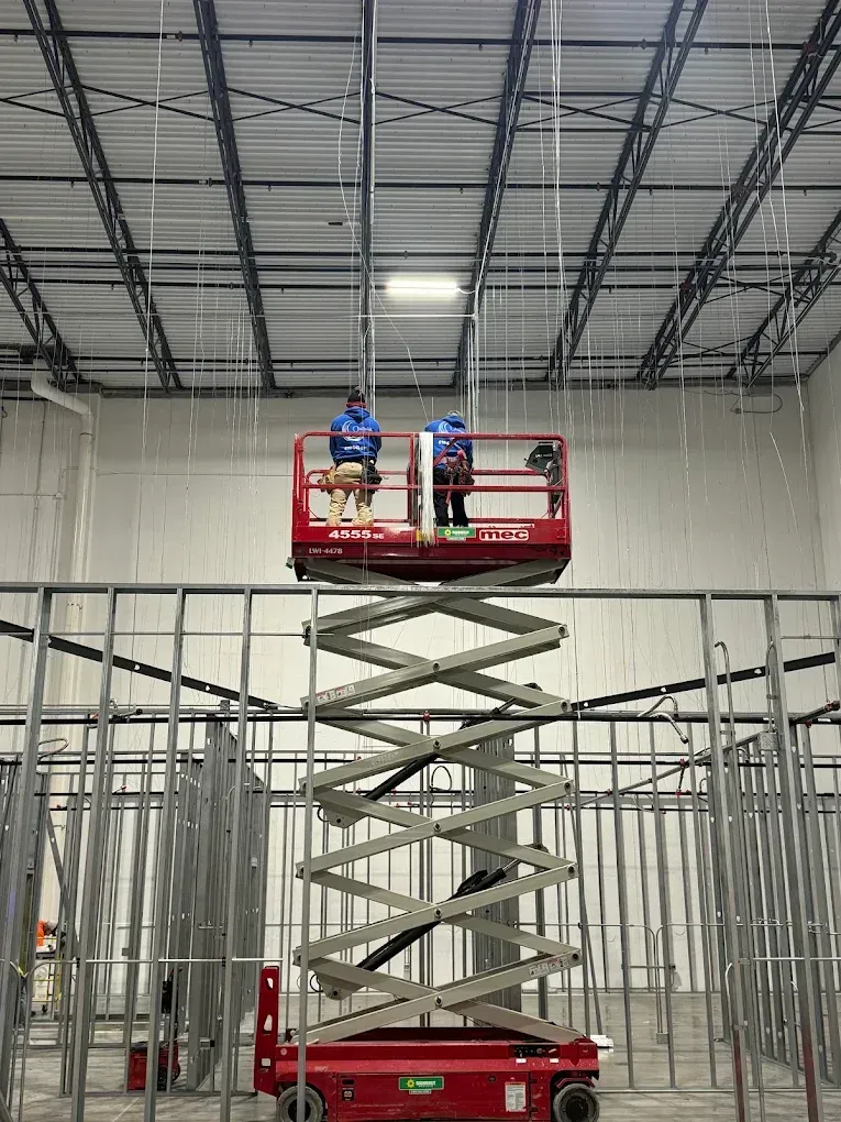 Two people on a red scissor lift install overhead wiring in a building under construction.