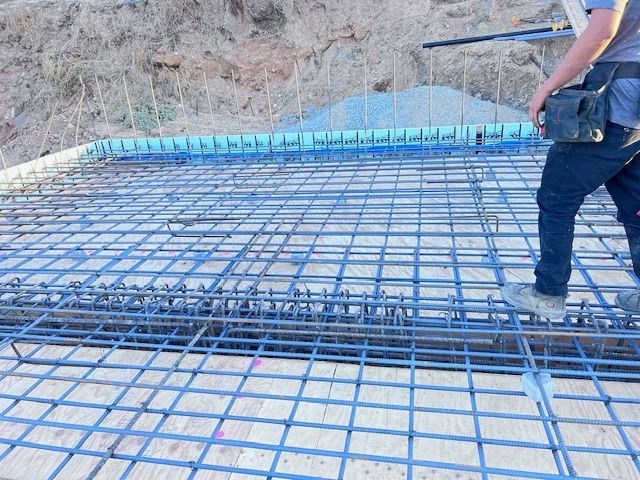 Construction worker standing over a blue rebar grid, preparing for concrete pouring. Outdoor setting.