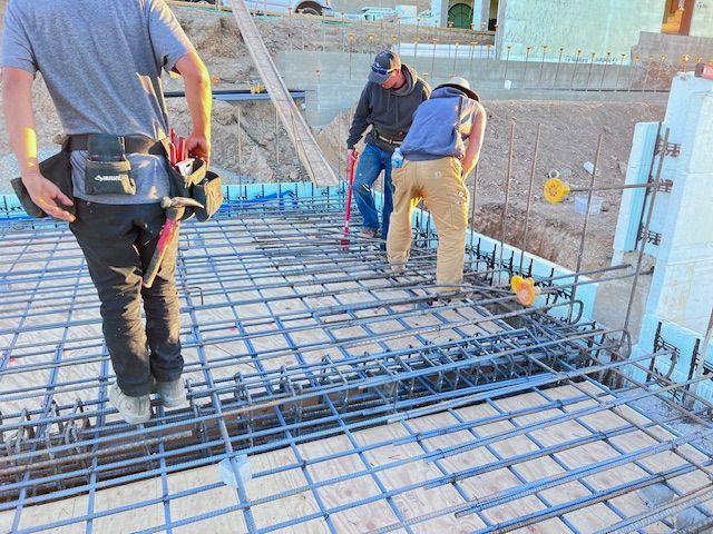 Construction workers on a foundation with rebar. Man in the foreground wearing a tool belt. Daylight.
