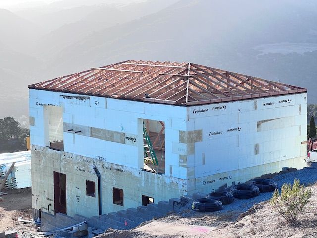 House under construction with light blue foam walls and wooden roof frame on a hillside.