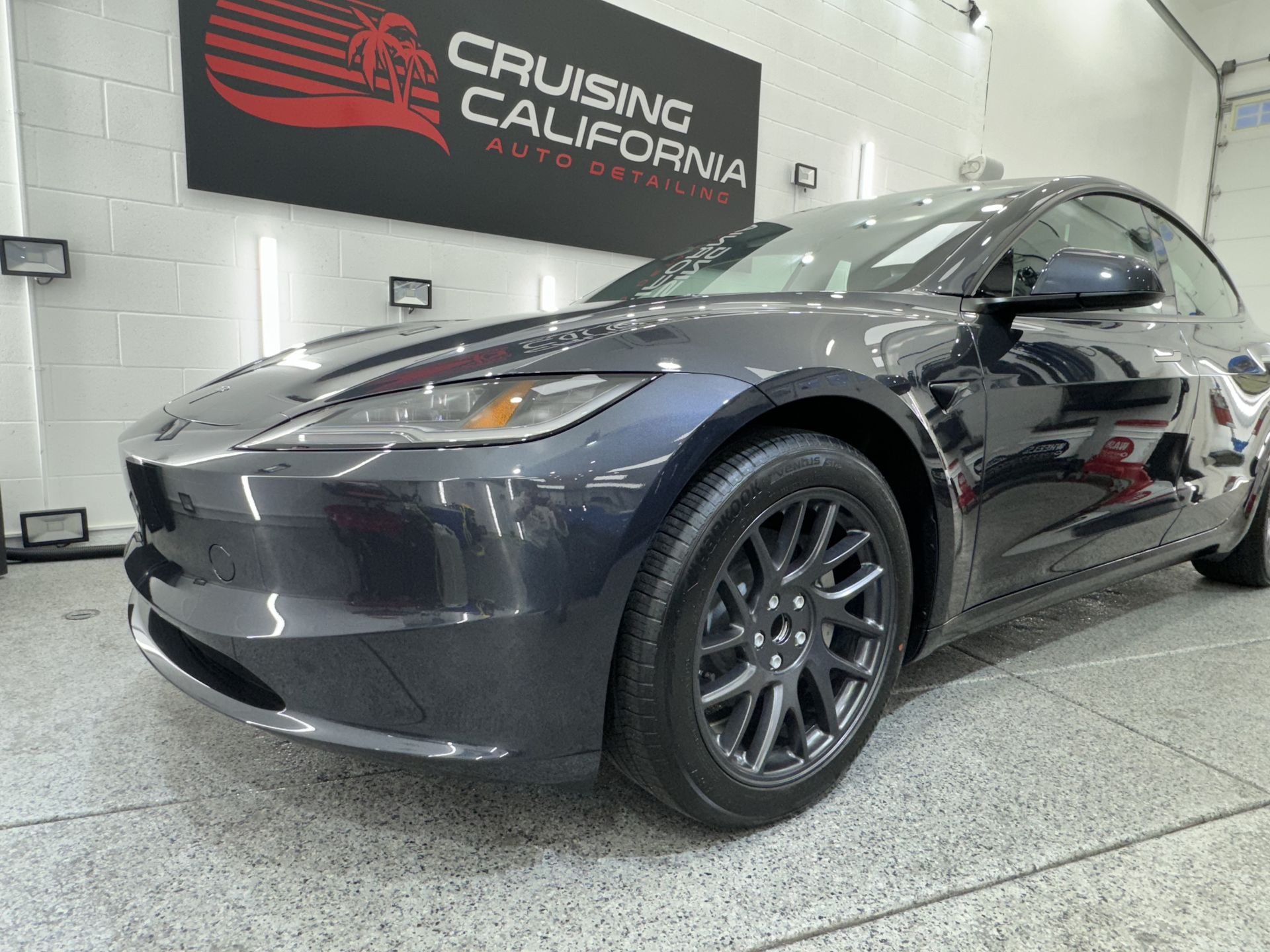 A black tesla model 3 is parked in front of a cruising california sign.