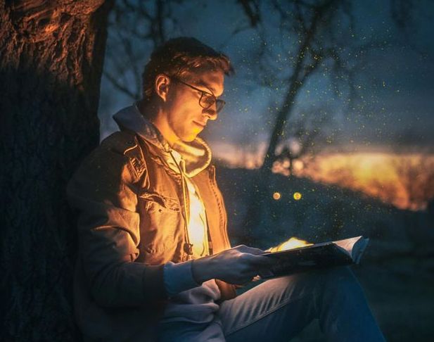 A man is sitting under a tree reading a book at night.