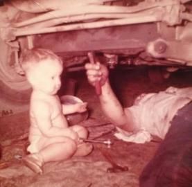 Child sits near person working on car undercarriage.