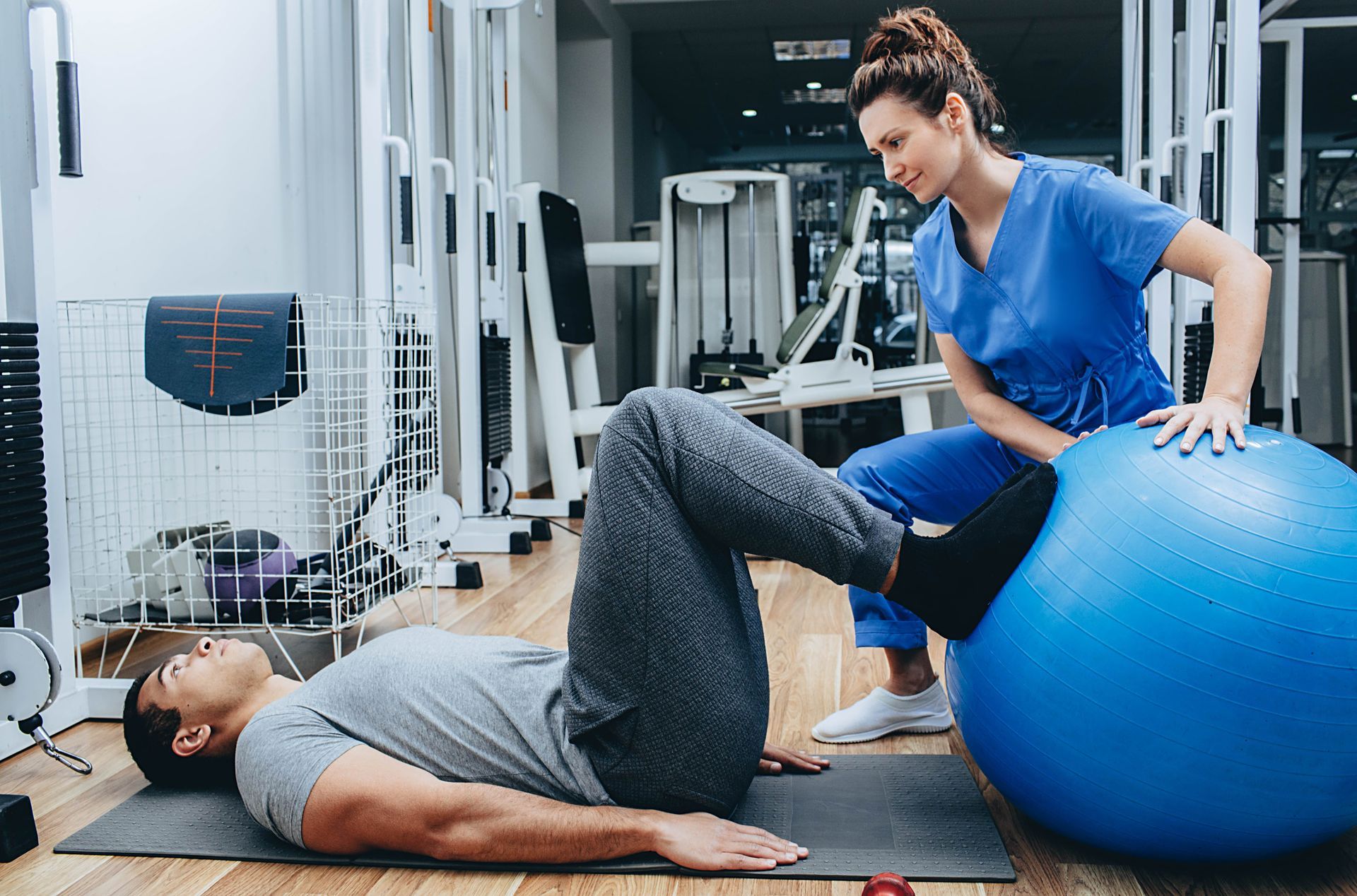 Person doing leg exercises on mat with stability ball and trainer in gym.