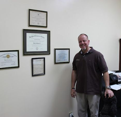 Man in brown shirt stands in front of a wall with framed documents. 